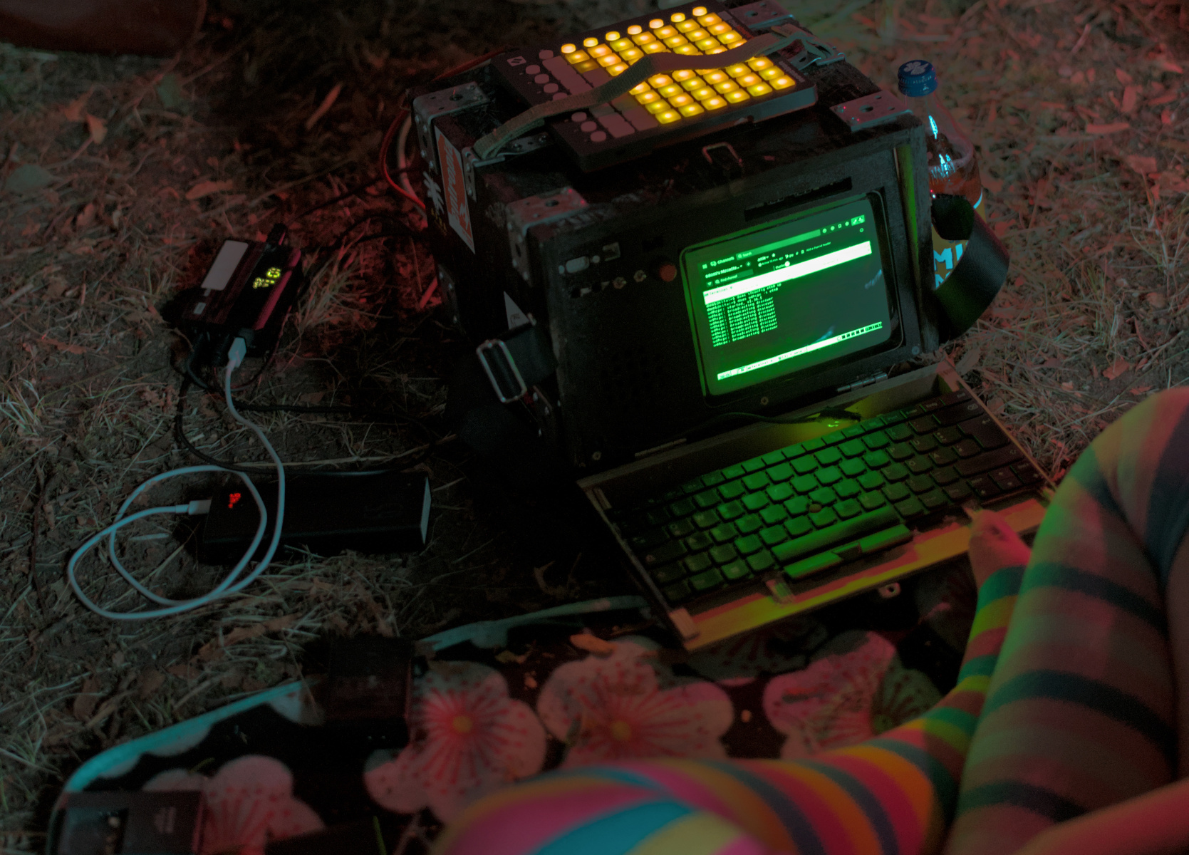 Picture of a black box on the ground. Right next to it is a power supply and a charging power bank; On top of it, there's a blinky implement with multiple lights. The box is opened, to reveal a keyboard, a green CRT and a bunch of switches. In front of the box, partially out of frame, sits the creator; you can only see pride knee-high socks (left is pansexual pride, right is transgender pride)