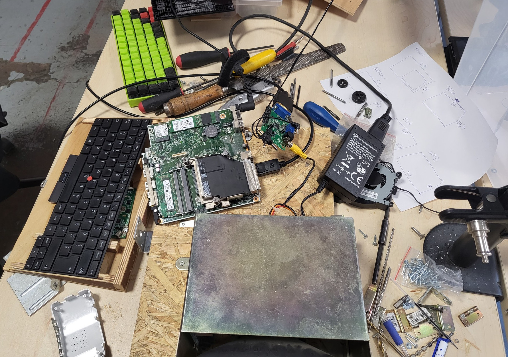 Picture of a desk with a ton of parts laying around, vaguely connected together. There's a keyboard on the left, next to it is a motherboard, next to that is a converter board from VGA to composite. You can also see a part of the display, next to a pile of hinges and brackets.